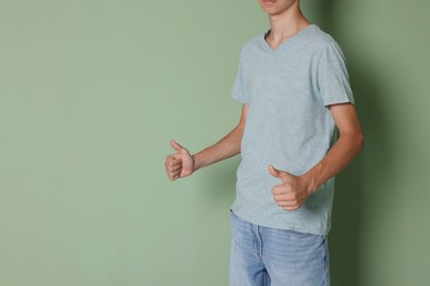 Photo of Teenage boy wearing light grey t-shirt and showing thumbs up on green background, closeup. Space for text