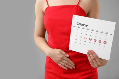 Woman holding calendar with marked menstrual cycle days and suffering from abdominal pain on grey background, closeup