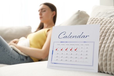 Photo of Woman using hot water bottle indoors, focus on calendar with marked menstrual cycle days on sofa