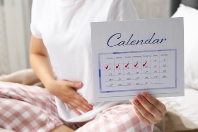 Photo of Woman holding calendar with marked menstrual cycle days on bed at home, closeup