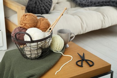 Basket with yarns, knitting needles, scissors and sweater on coffee table indoors