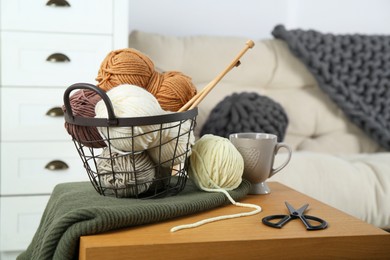 Photo of Basket with yarns, knitting needles, scissors and sweater on coffee table indoors