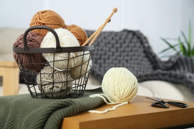 Photo of Basket with yarns, knitting needles, scissors and sweater on wooden table indoors