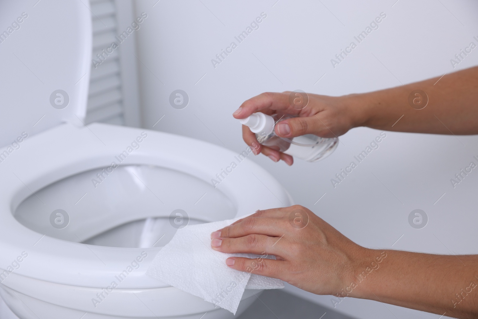 Photo of Woman cleaning toilet seat with paper and spray in bathroom, closeup