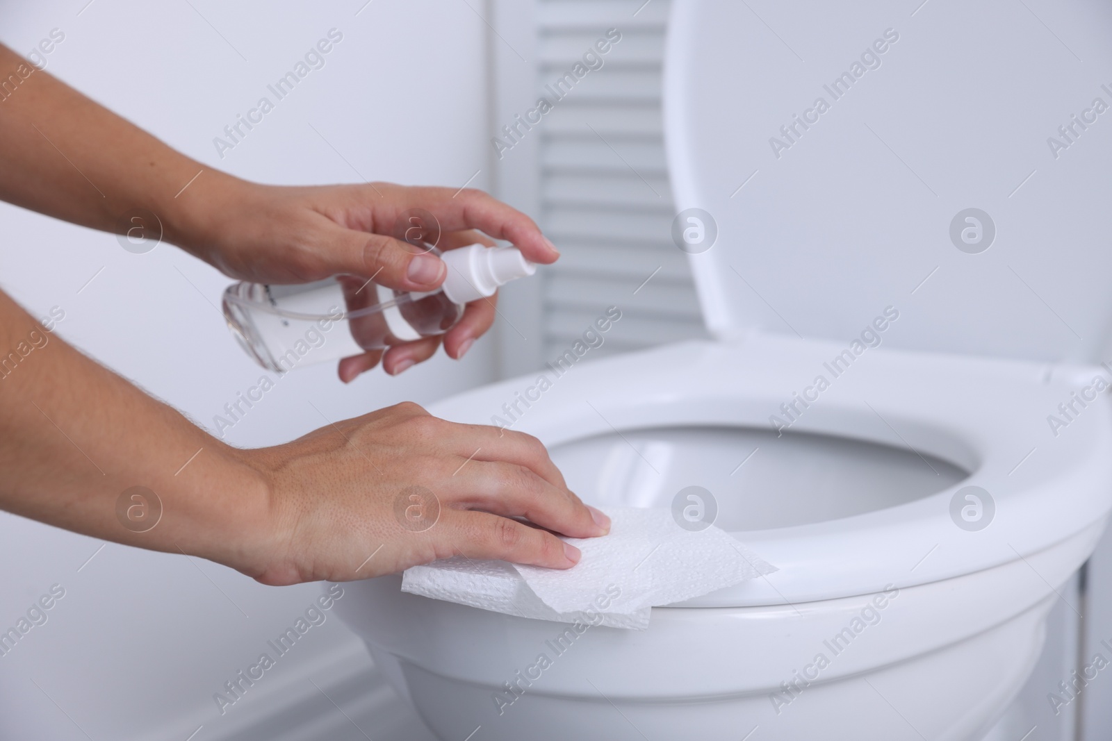 Photo of Woman cleaning toilet seat with paper and spray in bathroom, closeup