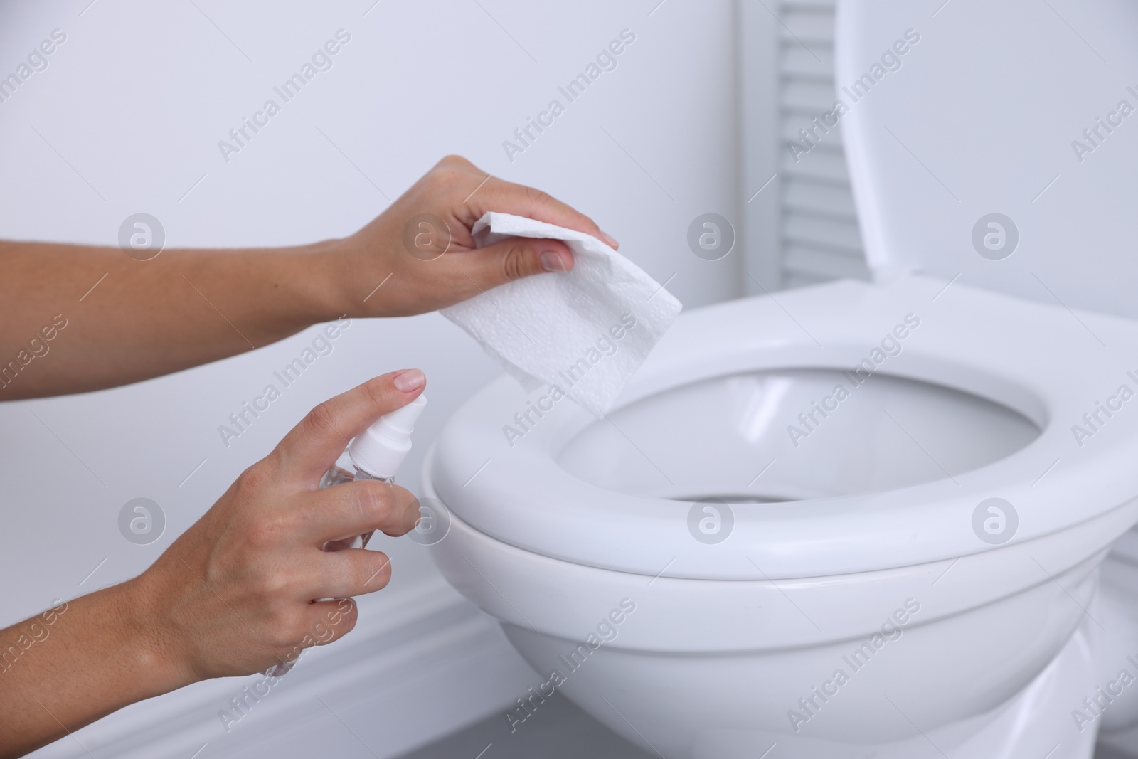 Photo of Woman cleaning toilet seat with paper and spray in bathroom, closeup