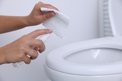 Woman cleaning toilet seat with paper and spray in bathroom, closeup