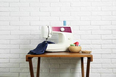 Photo of Sewing machine, blue fabric and spools of threads on wooden table near white brick wall