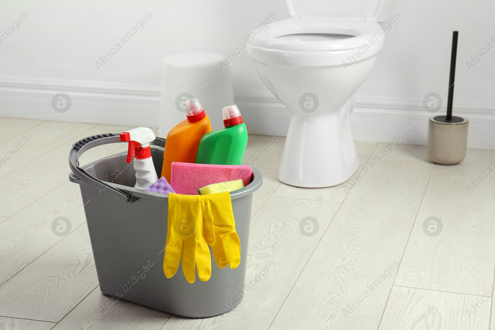 Photo of Bucket with different toilet cleaners, rag and gloves on floor in bathroom