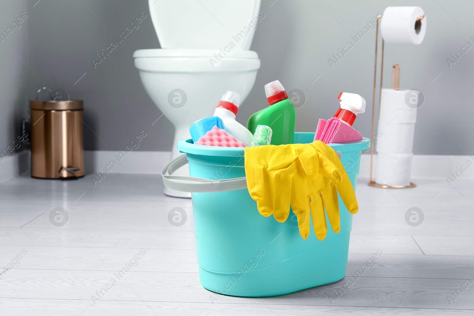 Photo of Bucket with different toilet cleaners, rag, sponge and gloves on floor in bathroom