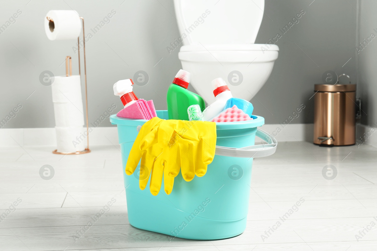 Photo of Bucket with different toilet cleaners, rag, sponge and gloves on floor in bathroom