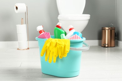 Photo of Bucket with different toilet cleaners, rag, sponge and gloves on floor in bathroom