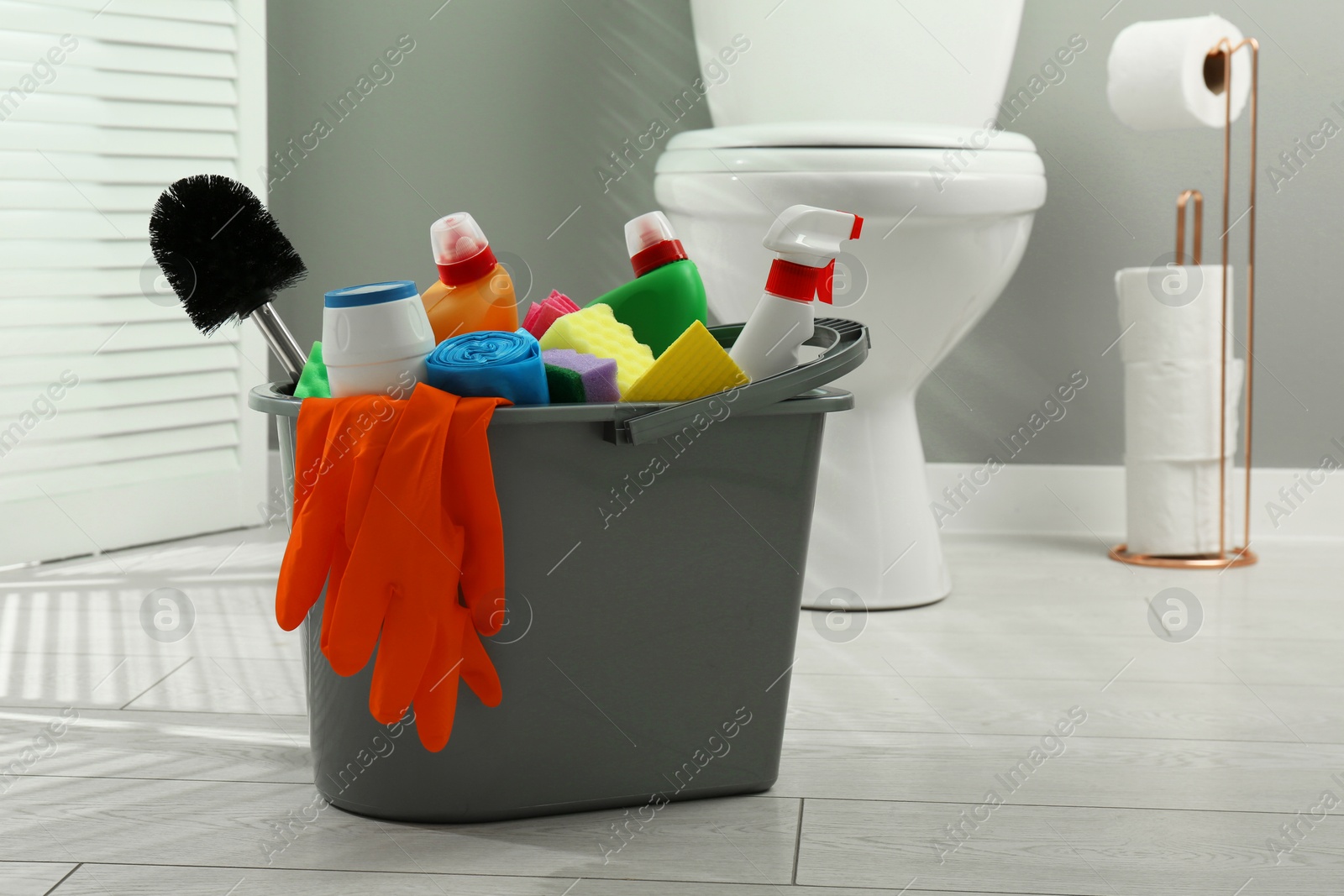 Photo of Bucket with different toilet cleaners, sponges, trash bags, brush and gloves on floor in bathroom