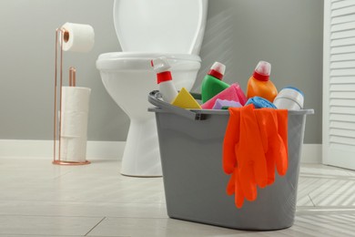 Photo of Bucket with different toilet cleaners, sponge and gloves on floor in bathroom