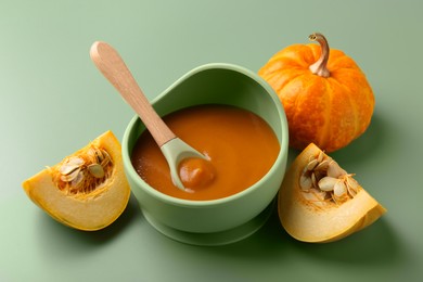 Photo of Delicious baby food with spoon in bowl and fresh pumpkins on green table