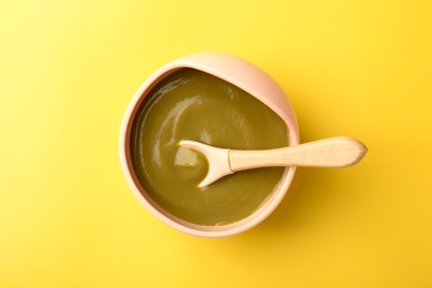 Photo of Delicious baby food with spoon in bowl on yellow table, top view