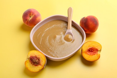 Photo of Delicious baby food with spoon in bowl and fresh peaches on yellow table