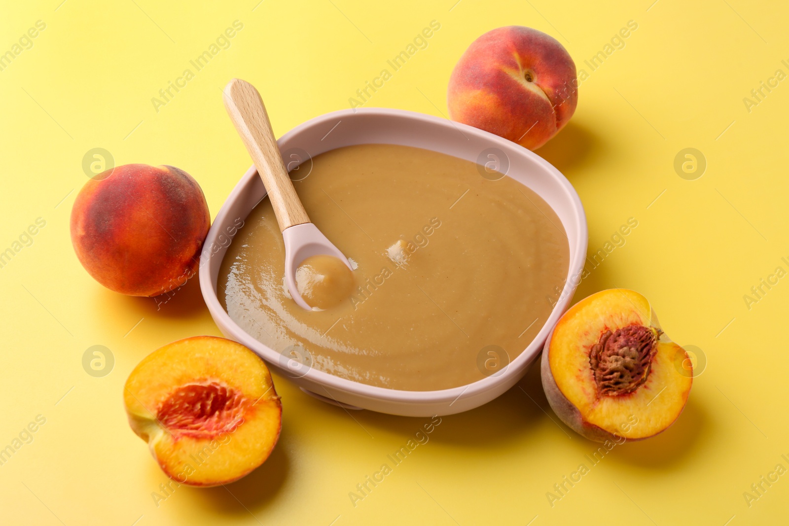 Photo of Delicious baby food with spoon in bowl and fresh peaches on yellow table