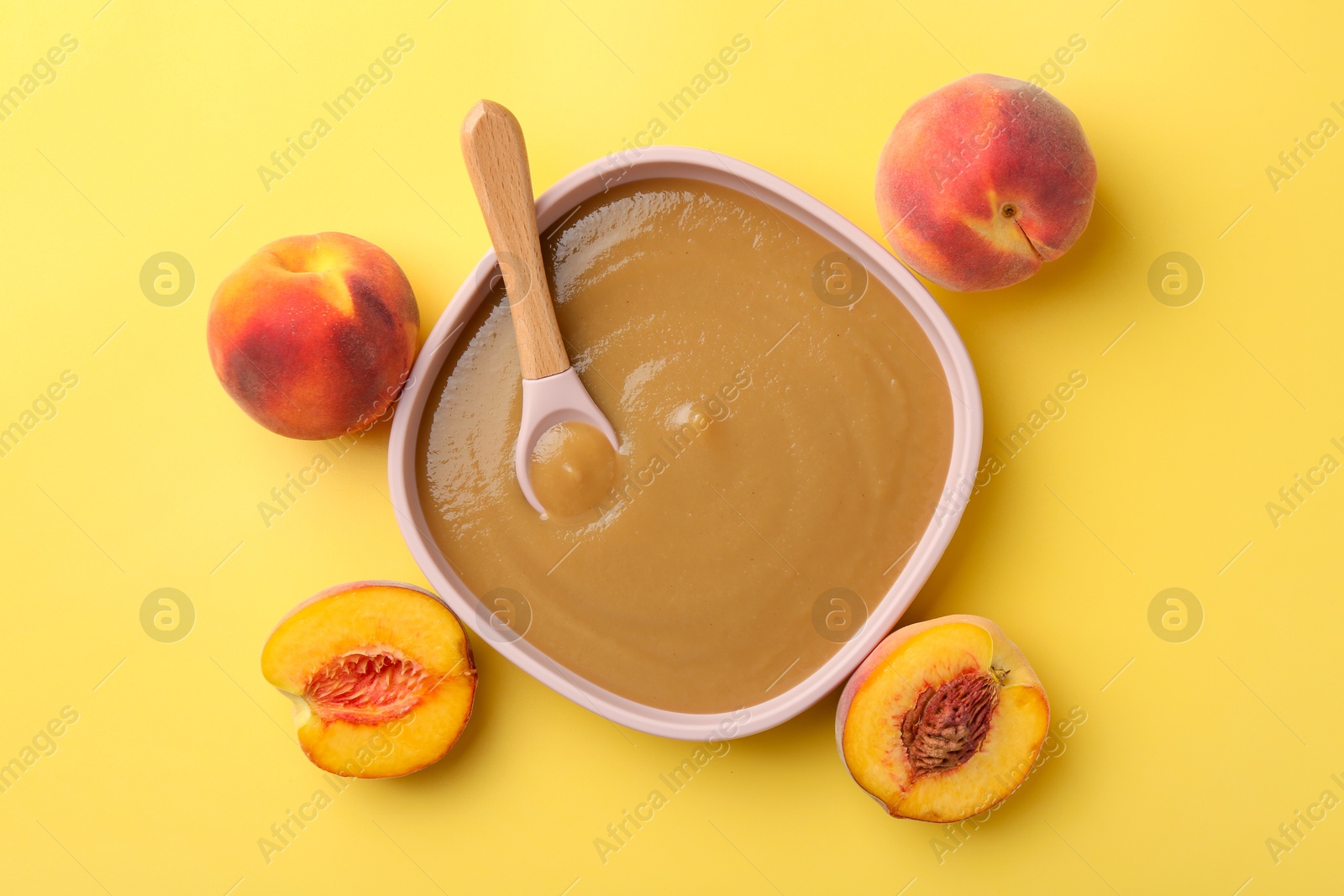 Photo of Delicious baby food with spoon in bowl and fresh peaches on yellow table, flat lay