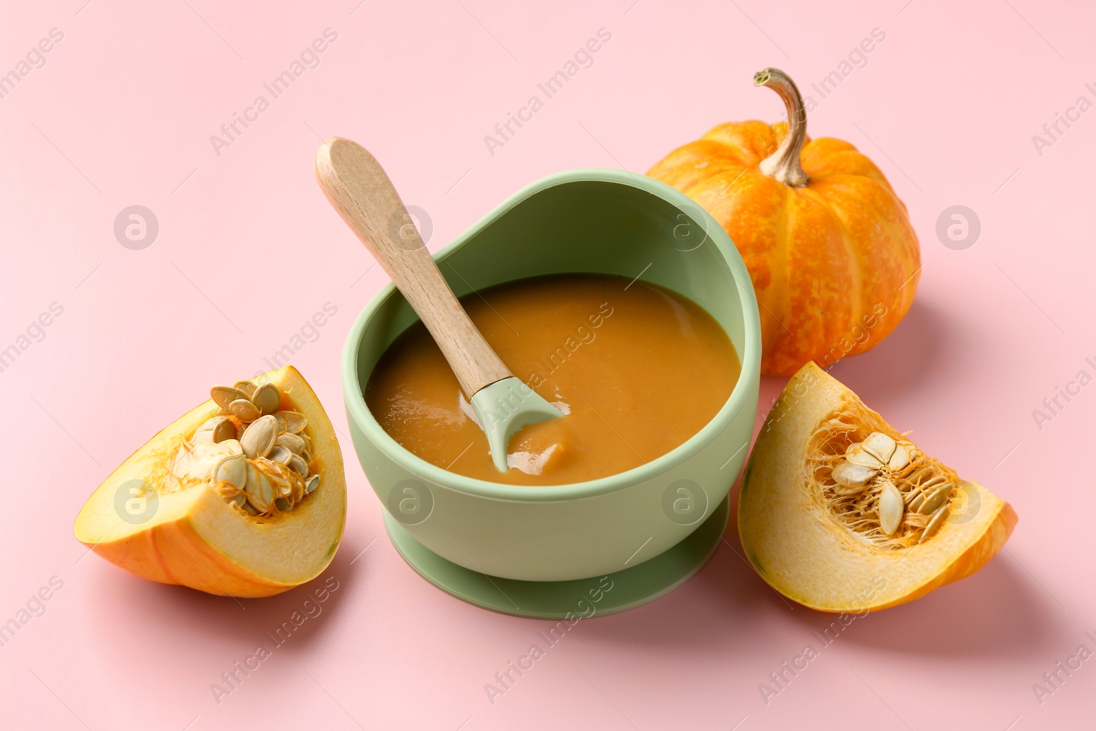 Photo of Delicious baby food with spoon in bowl and fresh pumpkins on pink table