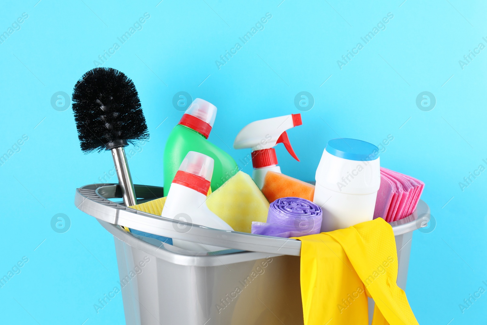 Photo of Bucket with different toilet cleaners, rags, rubber gloves and trash bags on light blue background