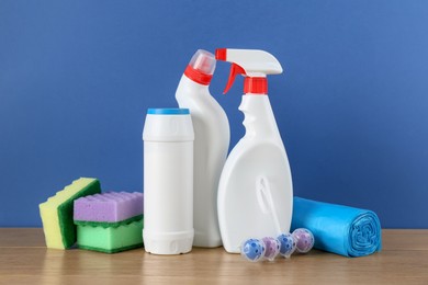 Photo of Different toilet cleaners, sponges and trash bags on wooden table against blue background