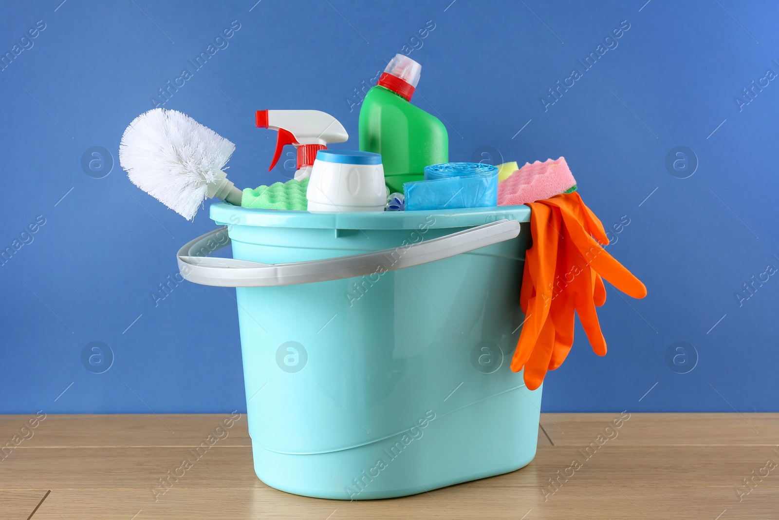 Photo of Bucket with different toilet cleaners, sponges, rubber gloves and trash bags on wooden table against blue background