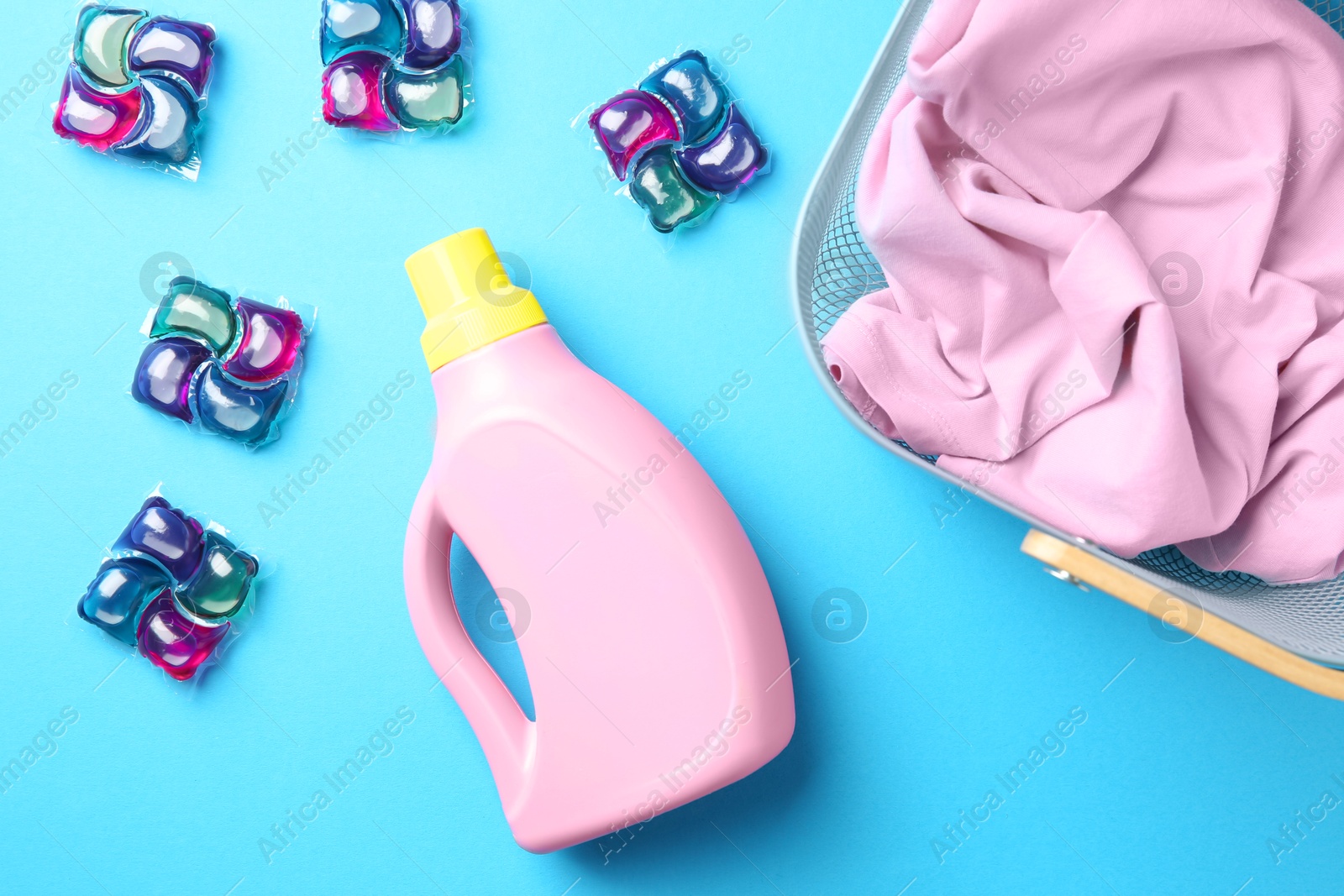 Photo of Basket with clothes, bottle of fabric softener and laundry detergent pods on blue background, flat lay