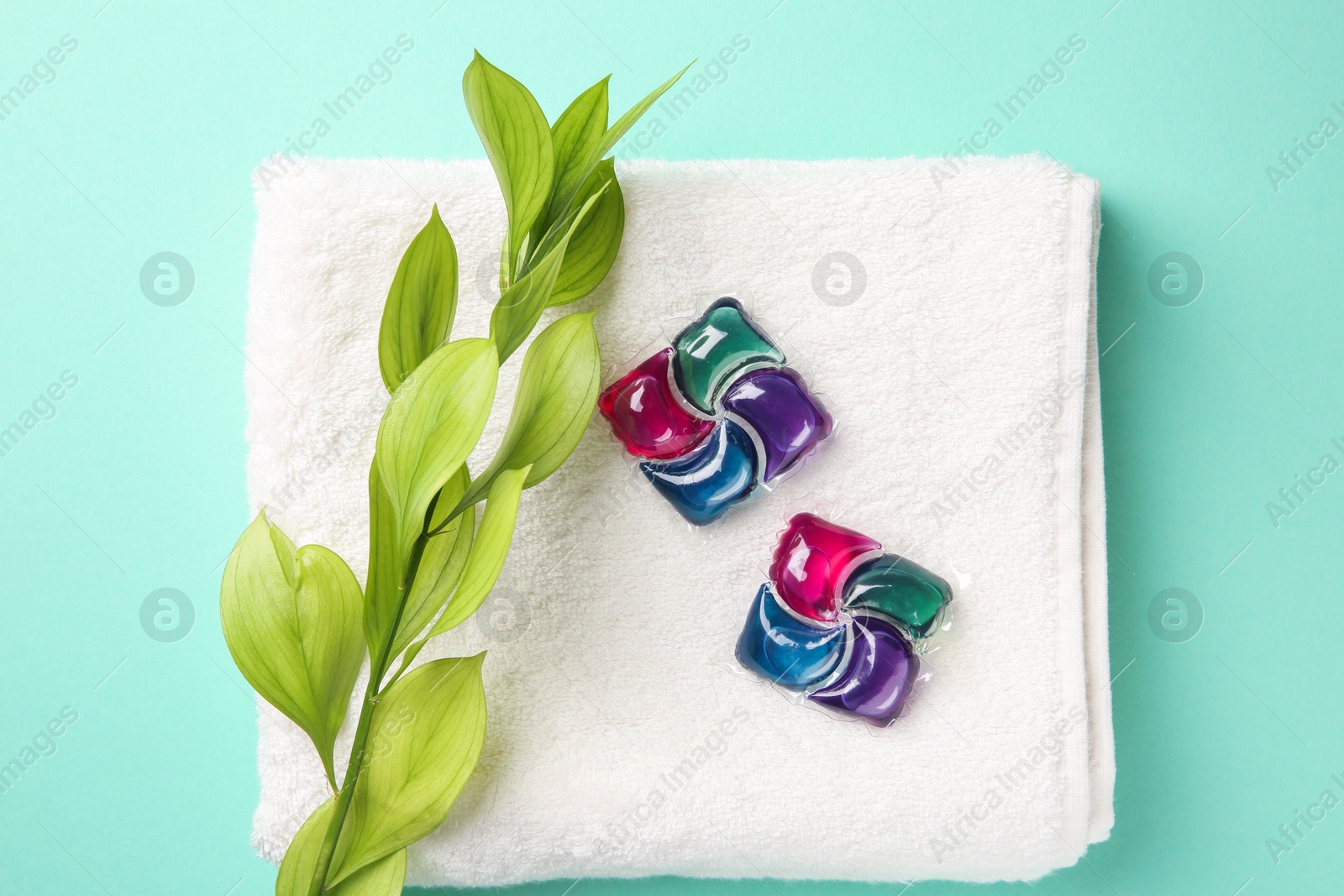 Photo of Laundry detergent pods, towel and green branch on turquoise background, flat lay