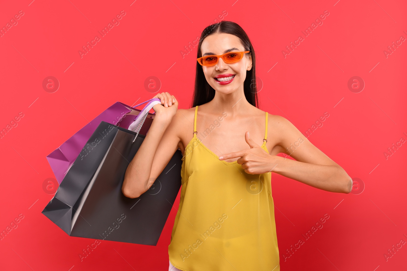 Photo of Smiling woman pointing at colorful shopping bags on red background