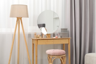 Mirror, dressing table, lamp and stool near window indoors. Interior design