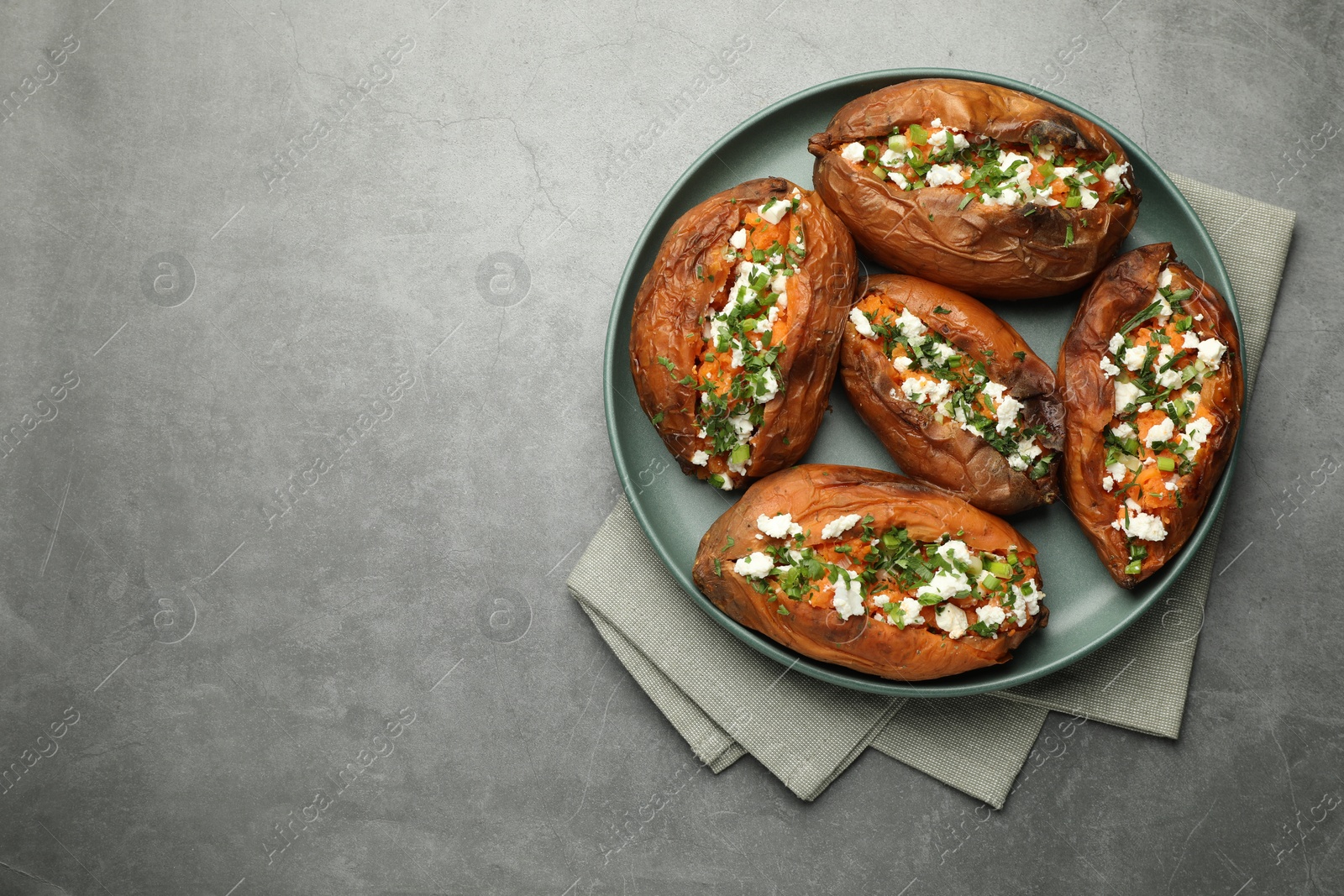 Photo of Tasty cooked sweet potatoes with feta cheese, green onion and parsley on gray textured table, top view. Space for text