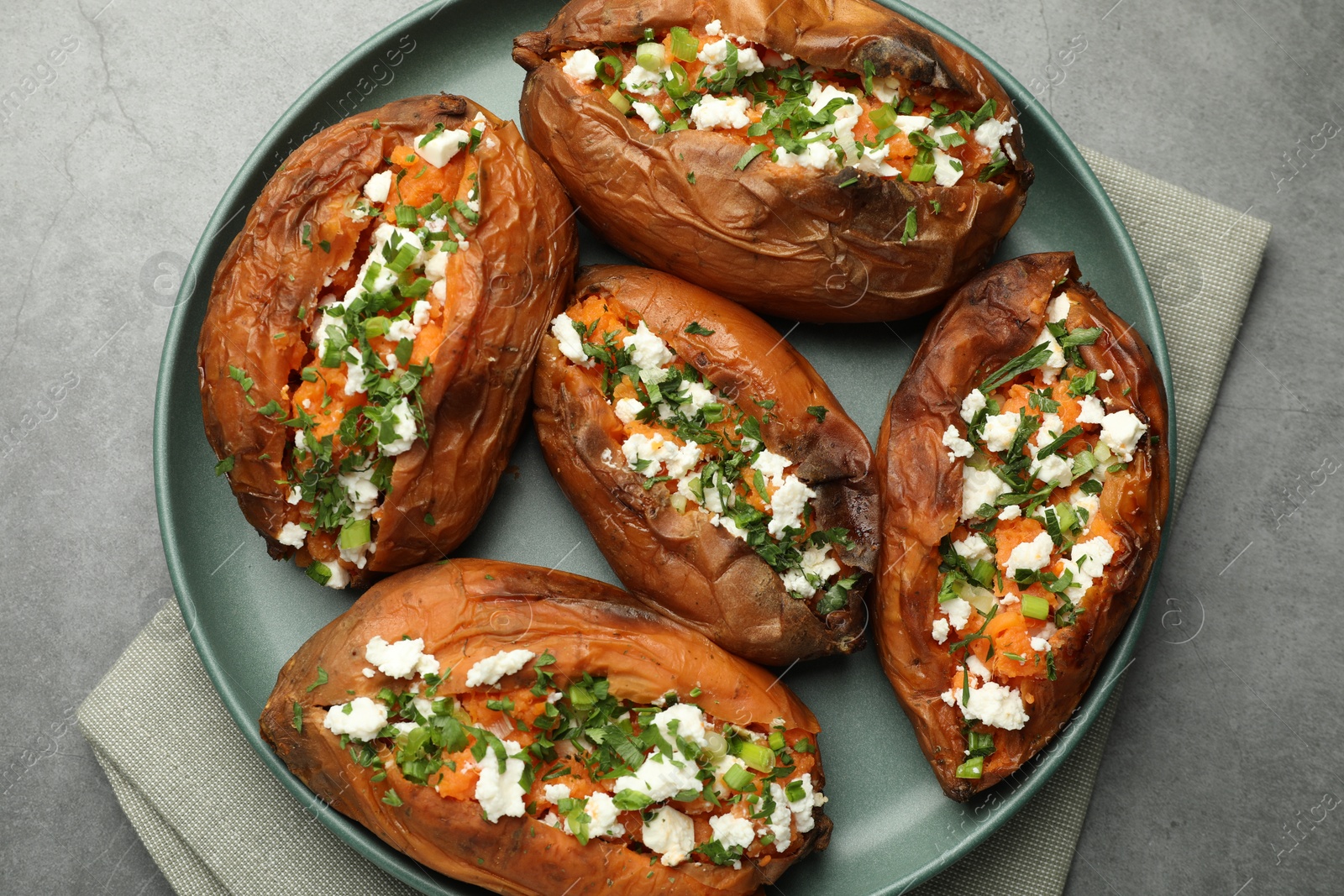 Photo of Tasty cooked sweet potatoes with feta cheese, green onion and parsley on gray textured table, top view