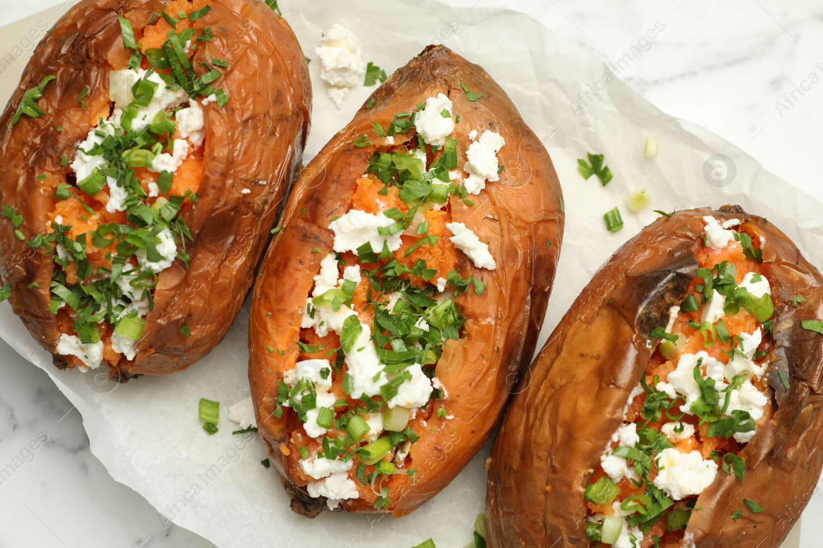 Photo of Tasty cooked sweet potatoes with feta cheese, green onion and parsley on white marble table, top view