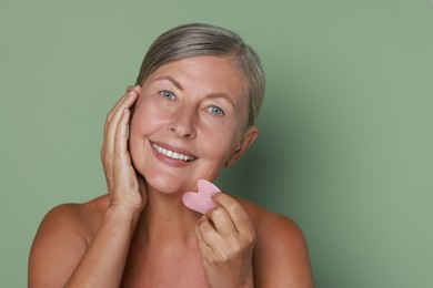 Beautiful young woman doing facial massage with gua sha tool on light green background