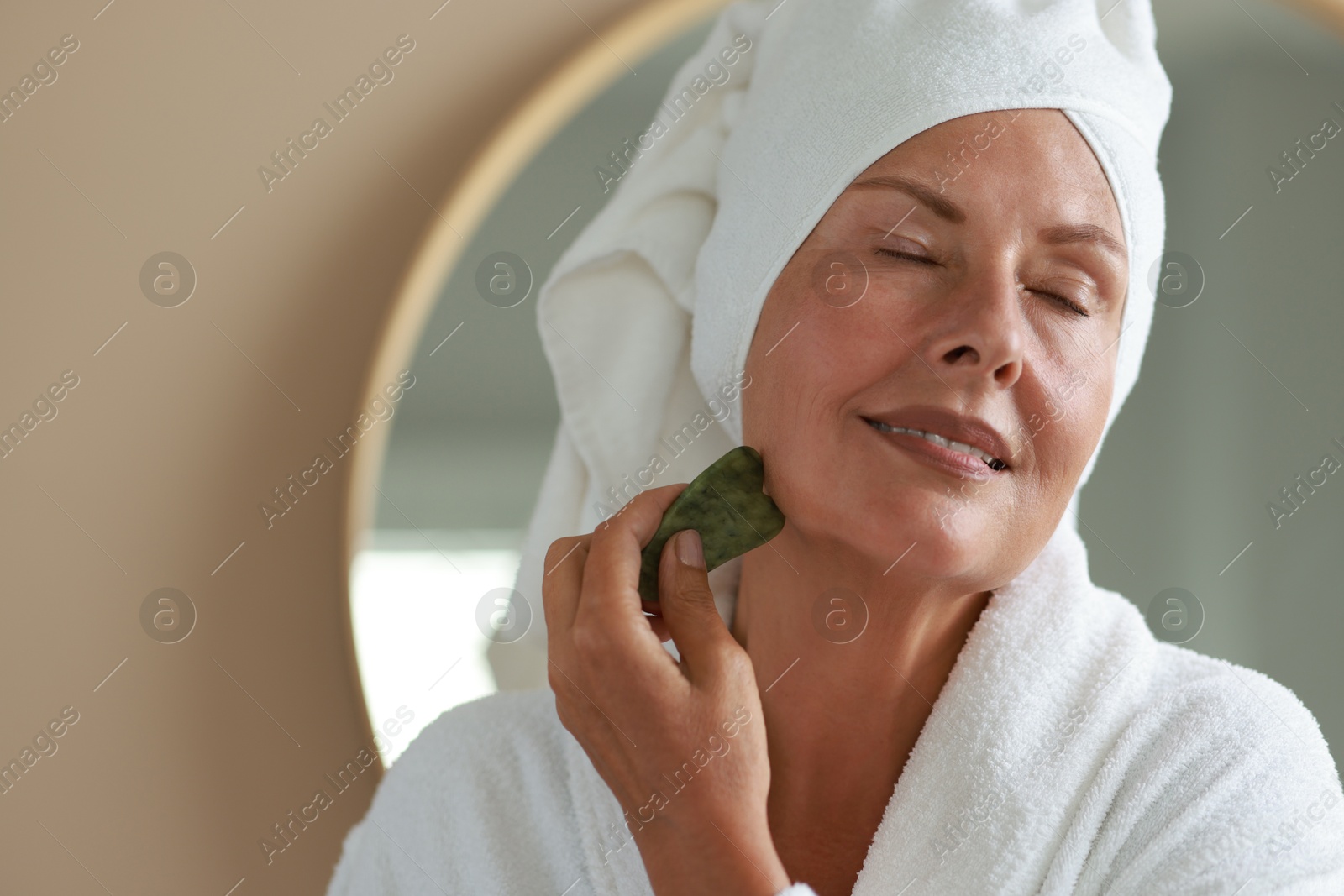 Photo of Beautiful young woman doing facial massage with gua sha tool indoors