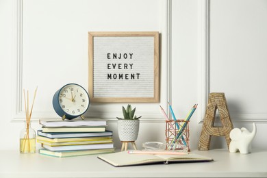 Photo of Letter board with phrase Enjoy Every Moment, alarm clock, decor and stationery on white desk