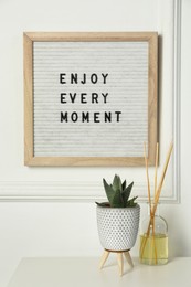 Photo of Letter board with phrase Enjoy Every Moment , reed diffuser and plant on white desk