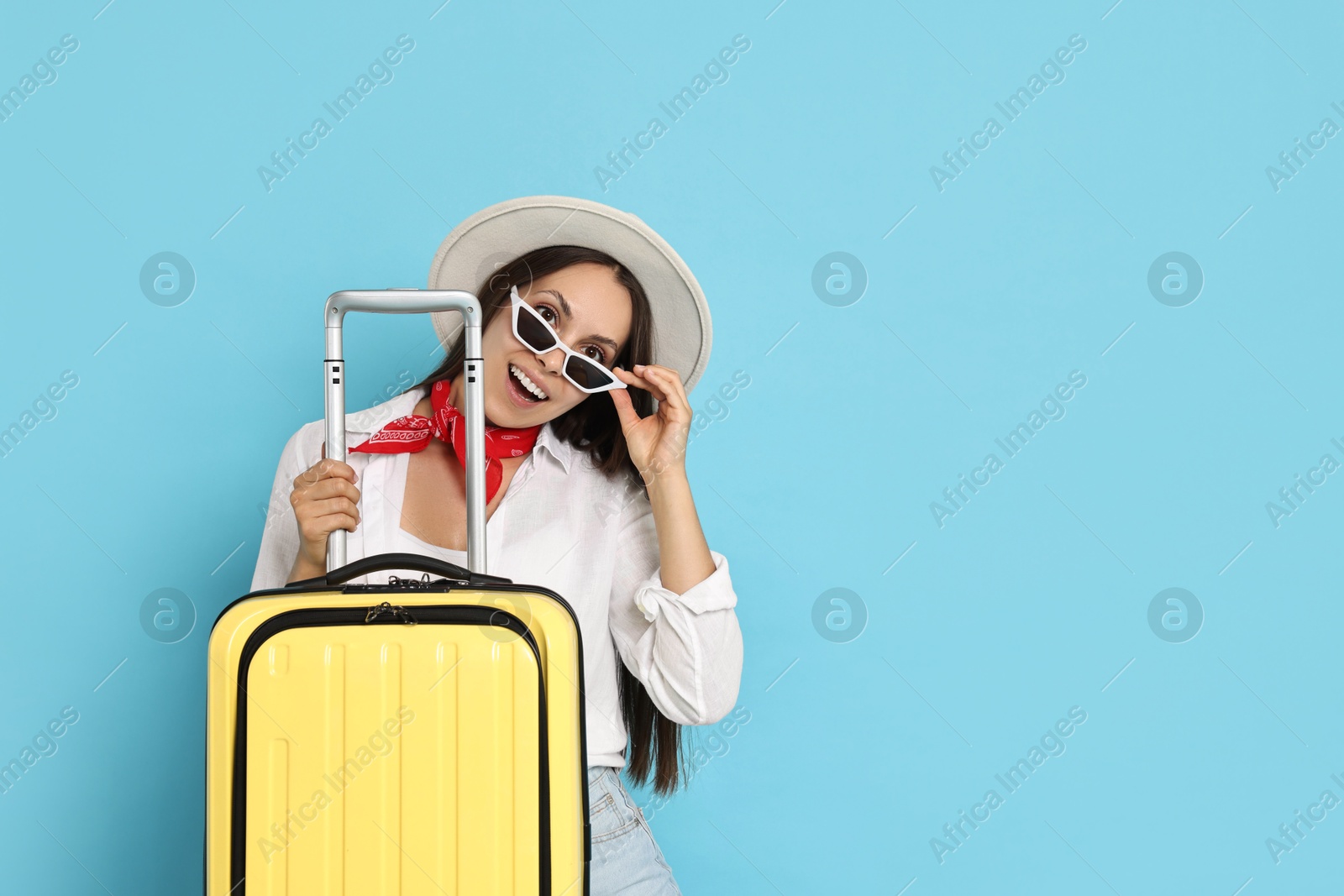 Photo of Happy young woman in sunglasses with suitcase and on light blue background, space for text