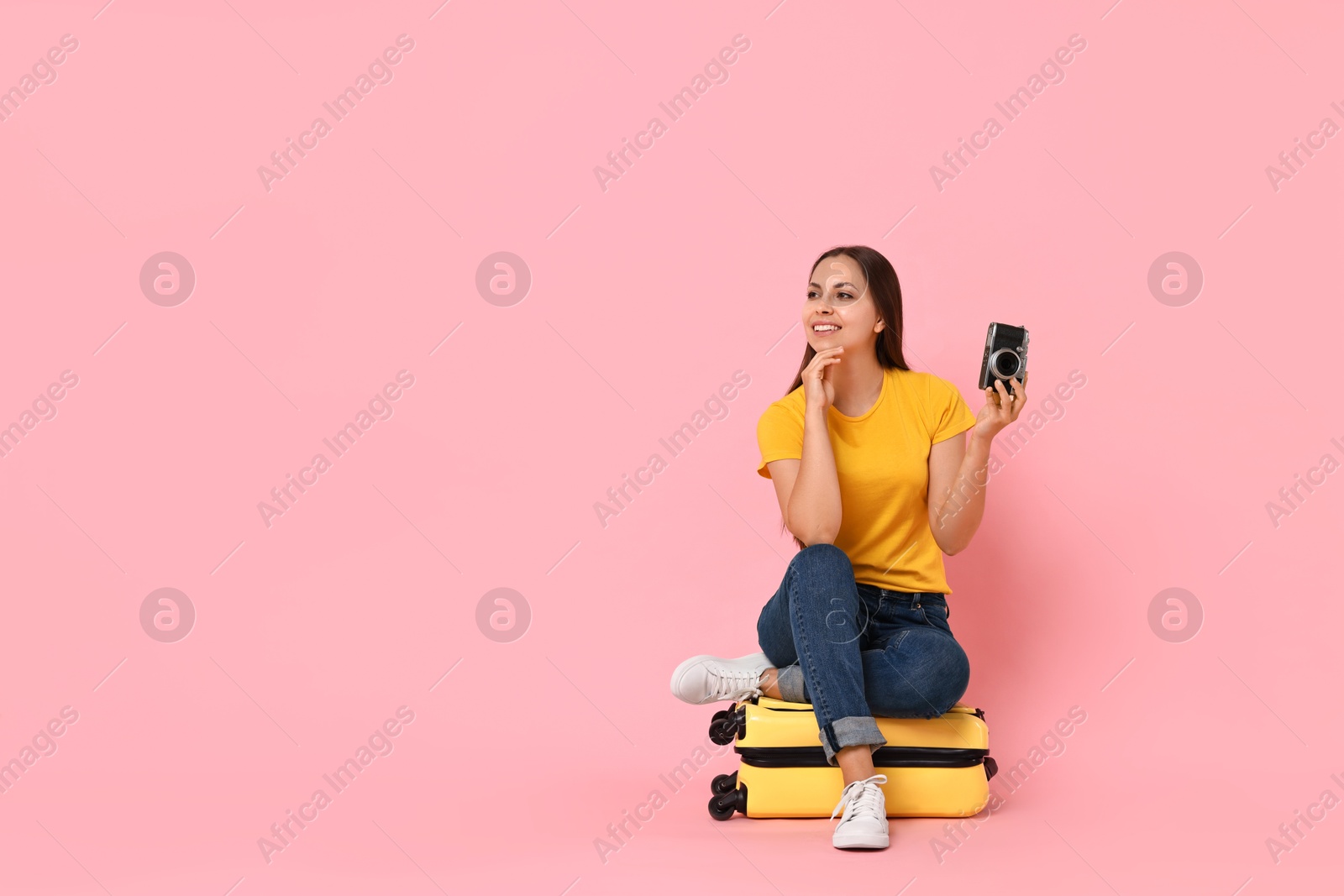 Photo of Happy young woman with vintage camera sitting on suitcase against pink background, space for text