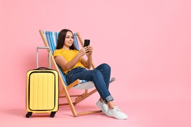 Happy young woman with smartphone sitting on folding chair and suitcase against pink background