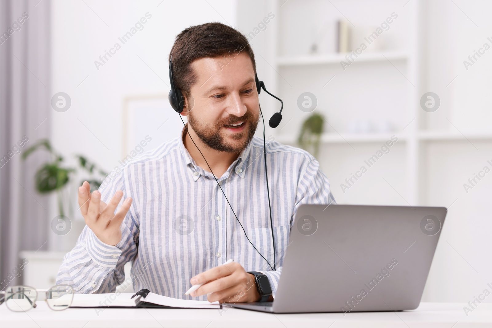 Photo of Interpreter in headset taking notes while having video chat via laptop at white table indoors