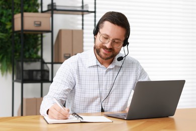 Interpreter in headset taking notes while having video chat via laptop at wooden table indoors