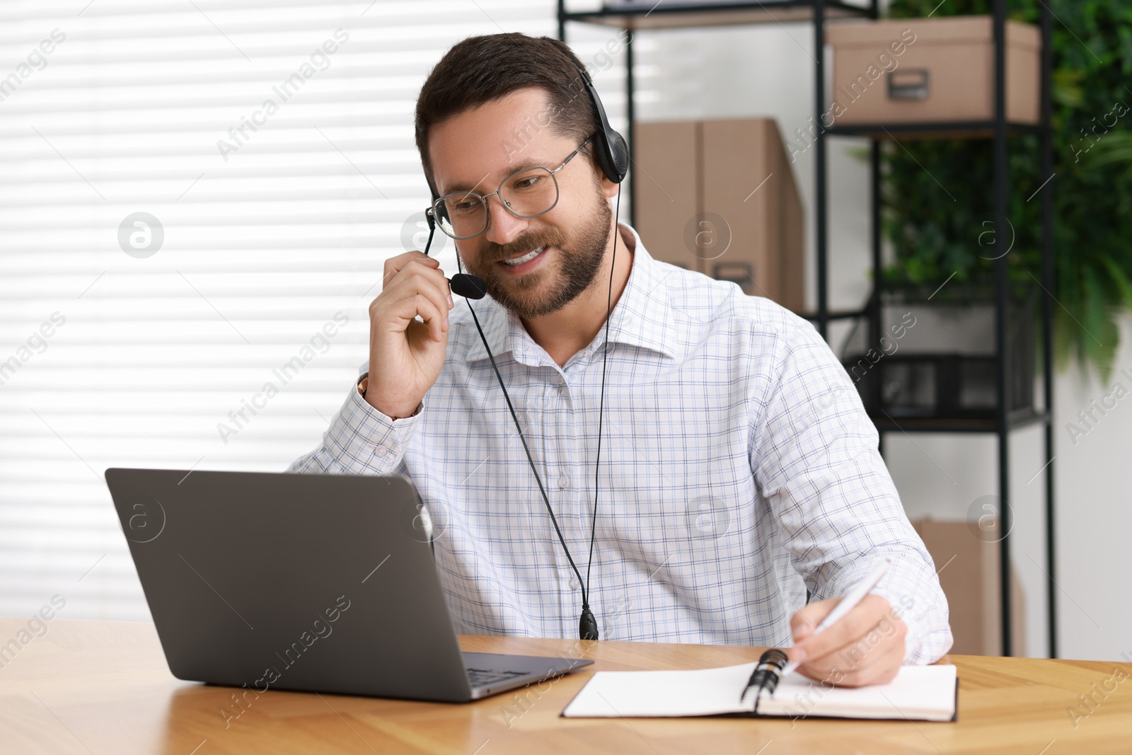 Photo of Interpreter in headset taking notes while having video chat via laptop at wooden table indoors