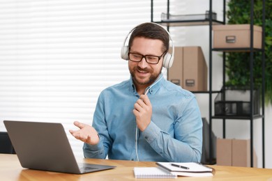 Photo of Interpreter in headphones having video chat via laptop at wooden table indoors