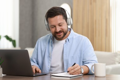 Photo of Interpreter in headphones taking notes while having video chat via laptop at wooden table indoors