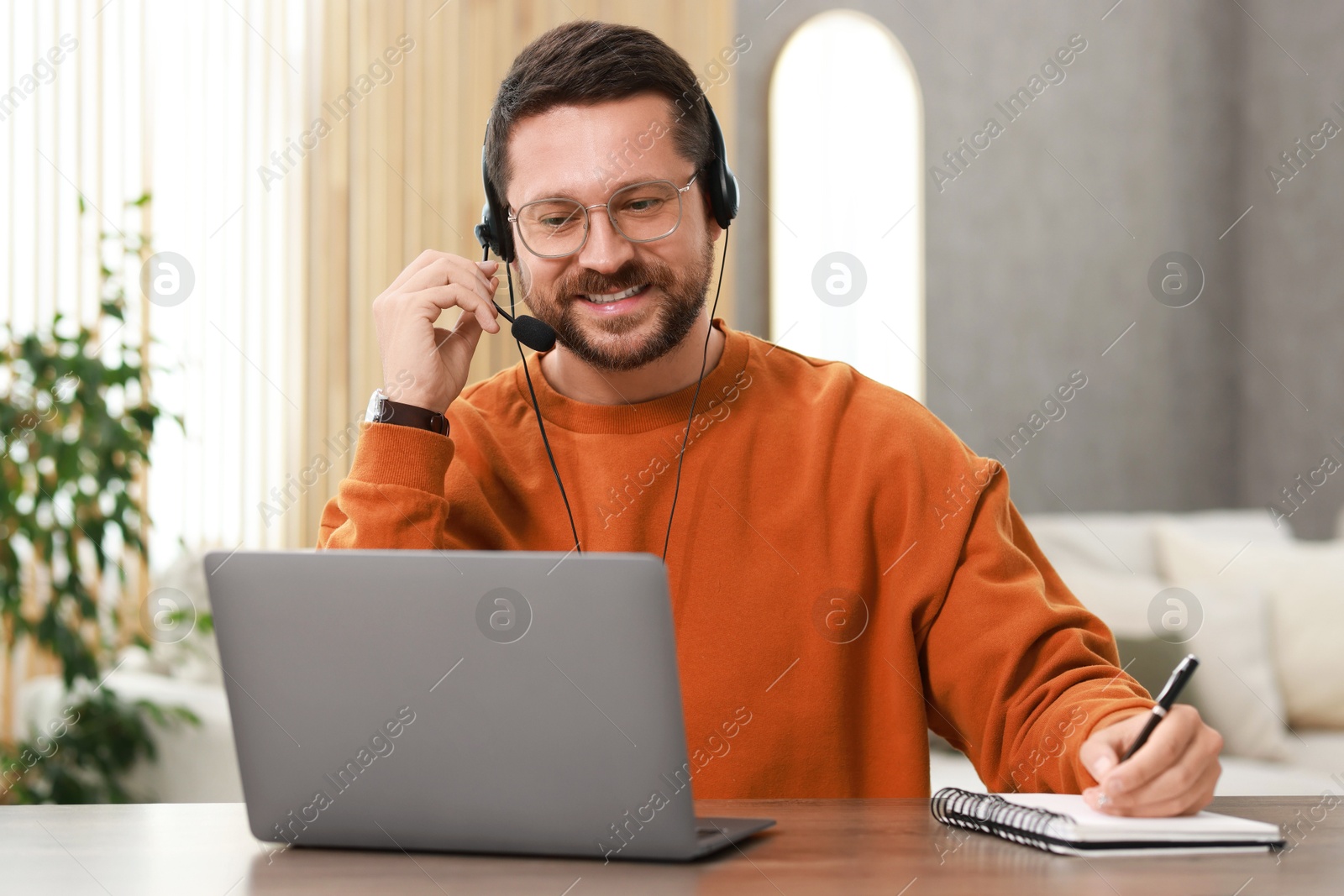 Photo of Interpreter in headset taking notes while having video chat via laptop at wooden table indoors