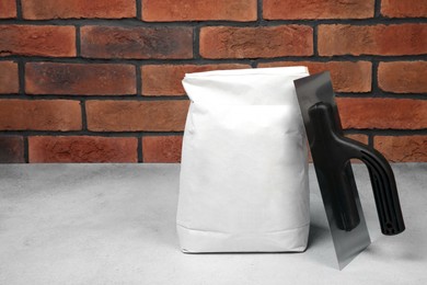 Bag of cement and plastering trowel on light textured table against red brick wall. Space for text