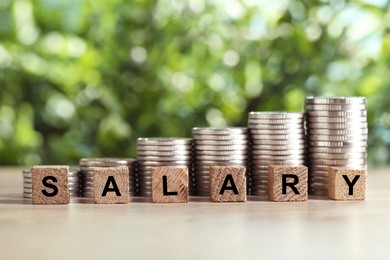 Photo of Word Salary made of cubes and stacked coins on light wooden table against blurred green background, closeup