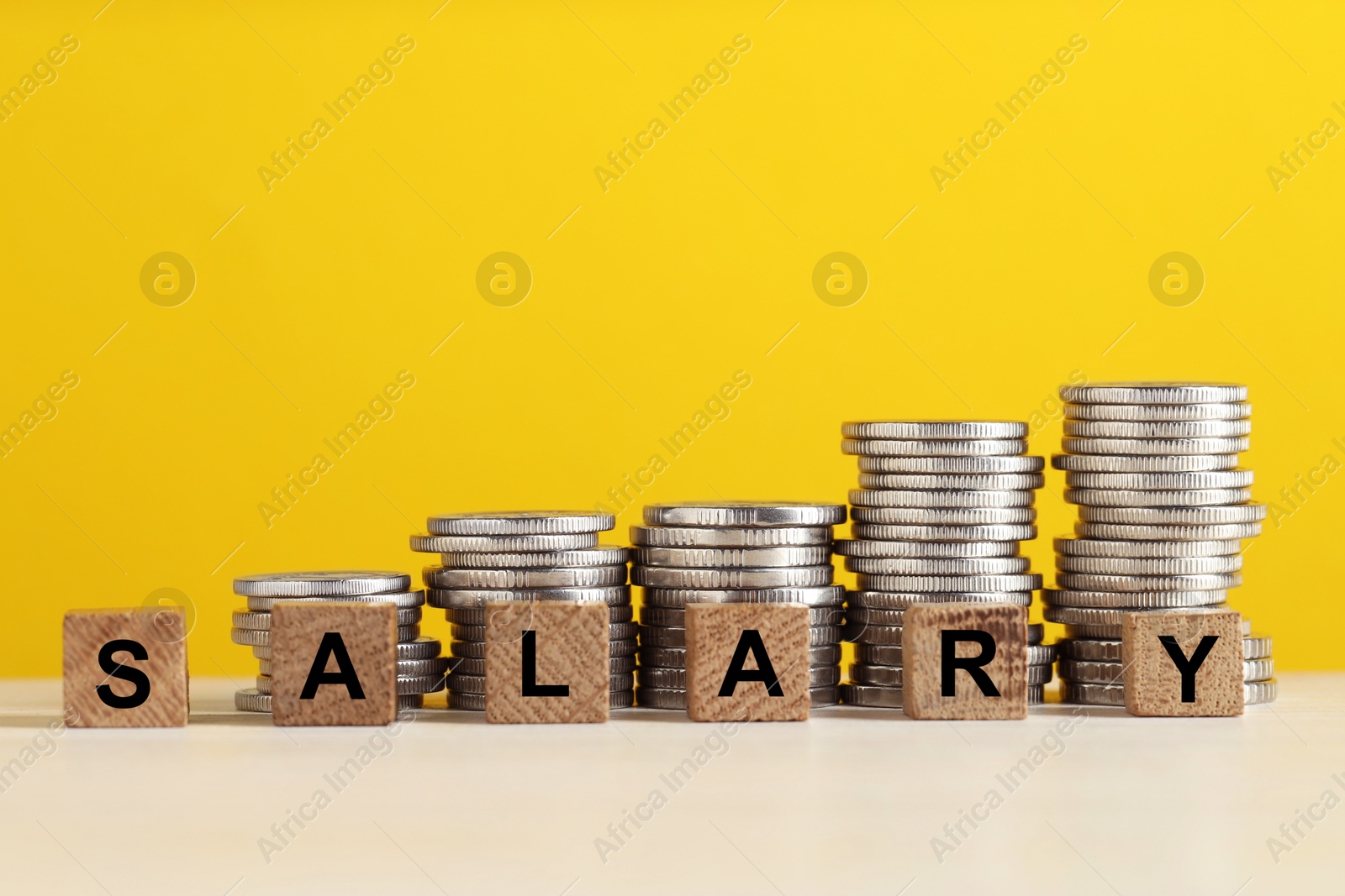 Photo of Word Salary made of cubes and stacked coins on white table against yellow background, closeup